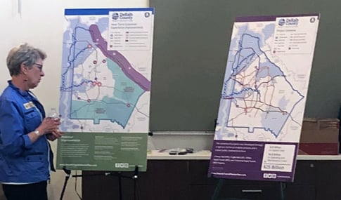 A woman stands next to two color renderings on easels of maps showing transit routes.