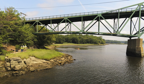 A group of men use an unmanned drone under a bridge crossing over a large river.