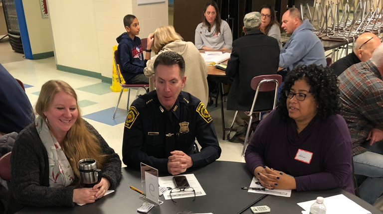 A diverse group of community members sit at a round table together while one person writes ideas on a large writing pad.