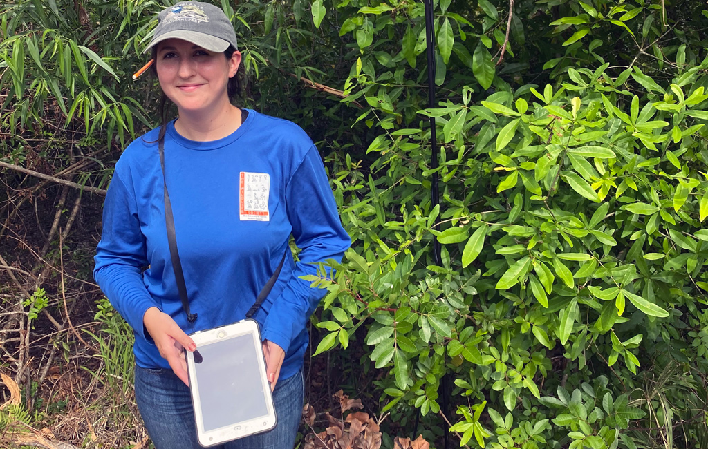 A woman in a long sleeve blue shirt, jeans, and ball cap holds an iPad and stands beside a pole used to collect acoustic data from bats