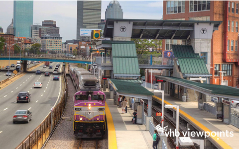 Commuters wait on a platform as a train pulls into the station, with the City of Boston in the background.