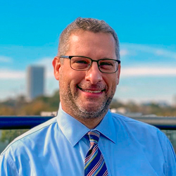 A business headshot of a man with brown hari wearing glasses, blue shirt and tie.